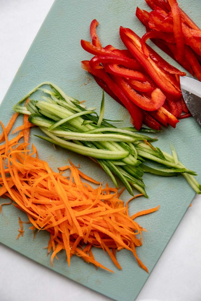 Shredded carrots, shredded cucumbers, and bell pepper on a blue cutting board.