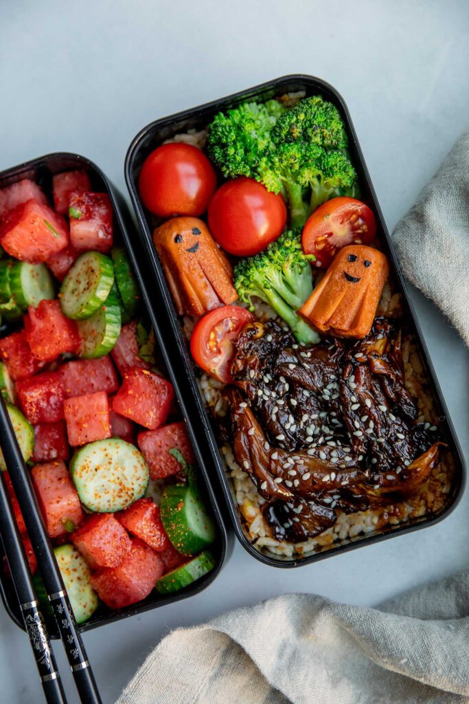 Side view of a designed plant based bento box. with watermelon salad in one container and a tempeh rice container topped with teriyaki mushrooms, steamed veggies and plant-based octopus sausage.