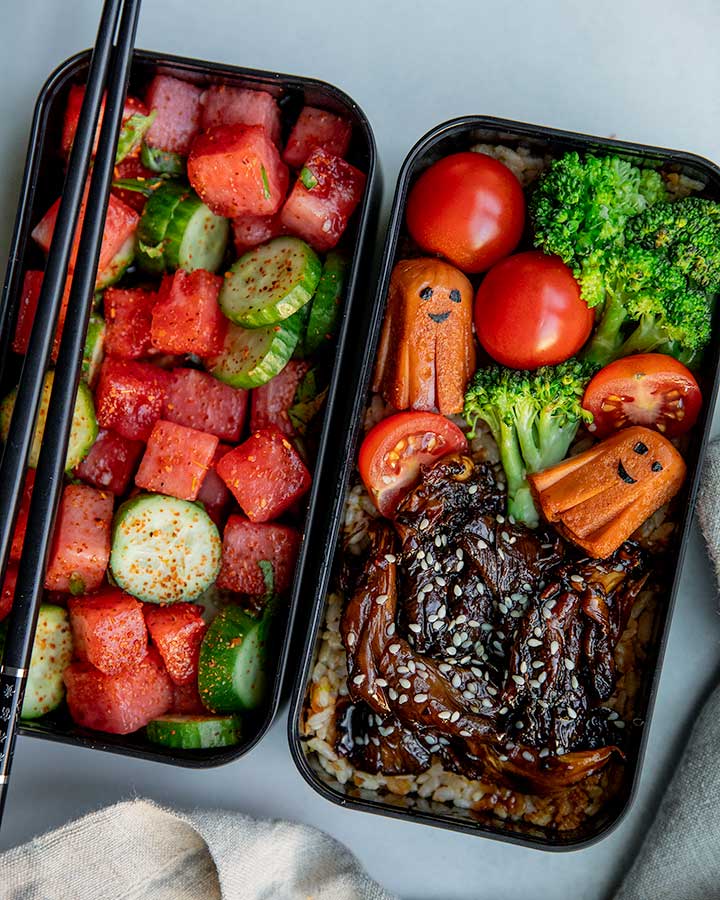 Assembled plant based bento with one compartment filled with watermelon salad and the second container filled with teriyaki mushrooms, high-protein tempeh rice, steamed veggies and plant based octopus sausage.
