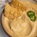 Bowl of cashew cheese sauce with two tortilla chips dug into the side and 2 slices of jalapeno.