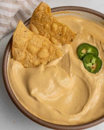 Bowl of cashew cheese sauce with two tortilla chips dug into the side and 2 slices of jalapeno.