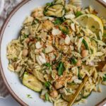 White bowl filled with a mixed zucchini orzo salad with sliced almonds, herbs and a wedge of lemon.