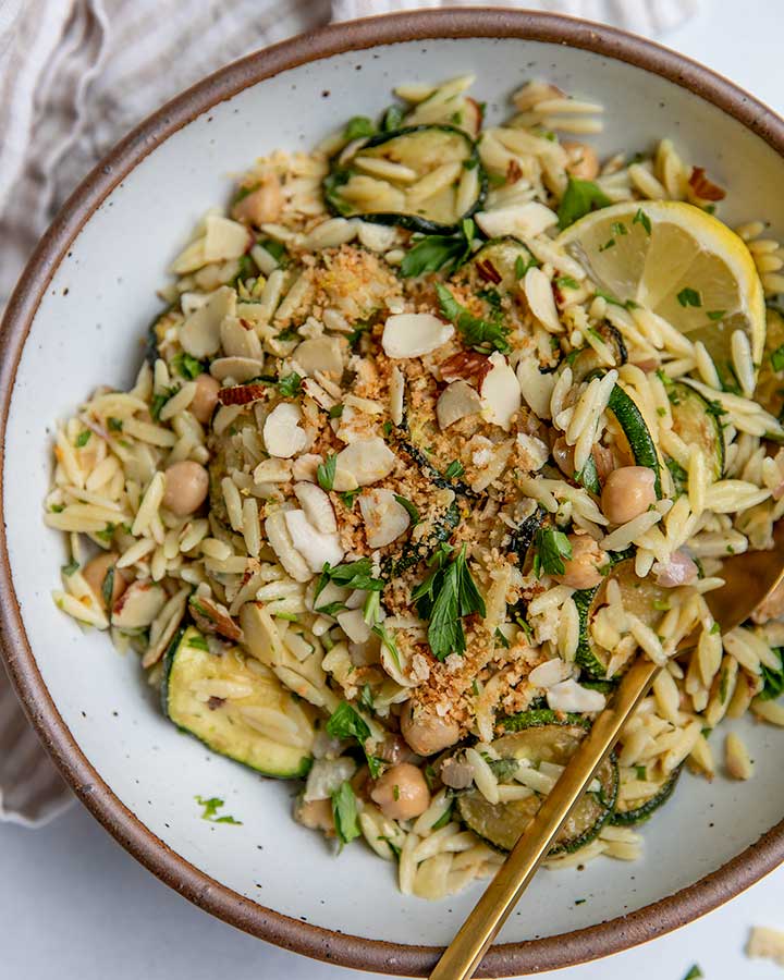 White bowl filled with a mixed zucchini orzo salad with sliced almonds, herbs and a wedge of lemon.