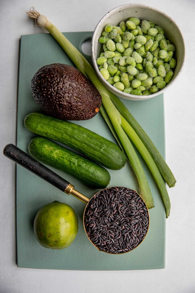 Avocado, scallions, bowl of edamame, 2 Persian cucumbers, cup of black rice and lime on a cutting board.
