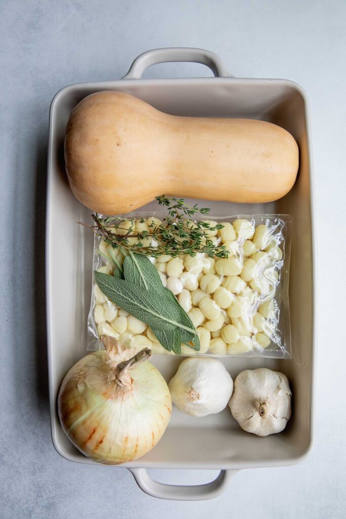 Baking dish filled with butternut squash, gnocchi, sage leaves, thyme, onion and garlic bulbs.