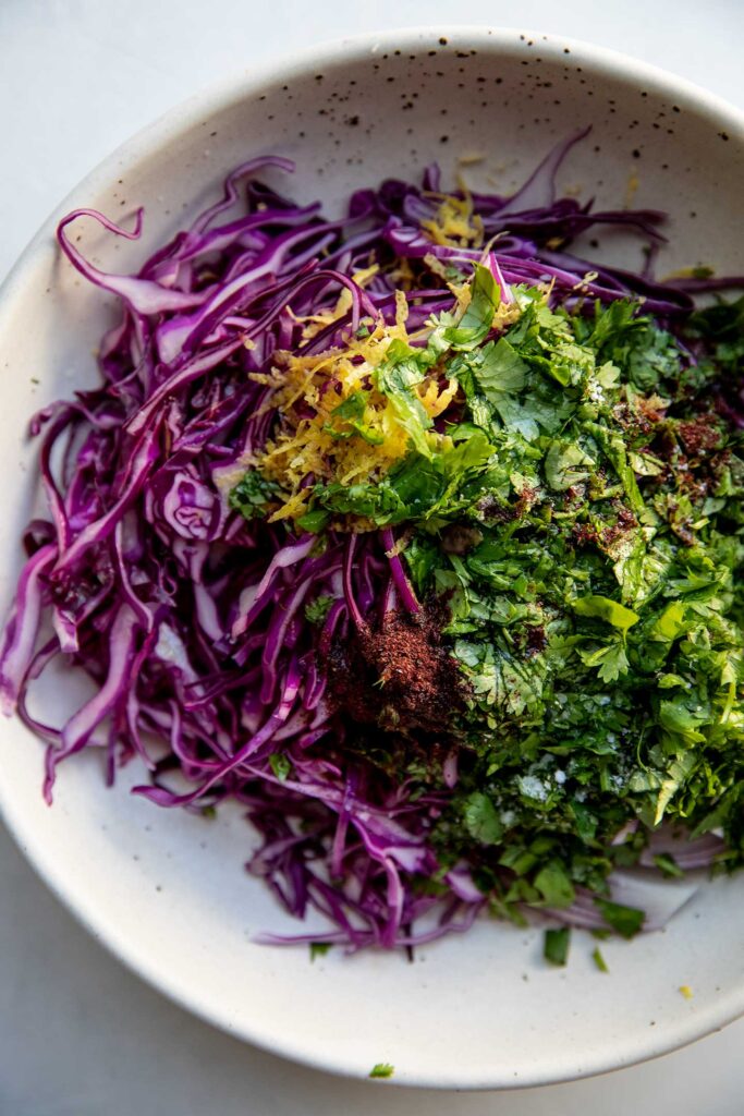 Cabbage, red onions, cilantro, lemon zest, and sumac in a white mixing bowl.