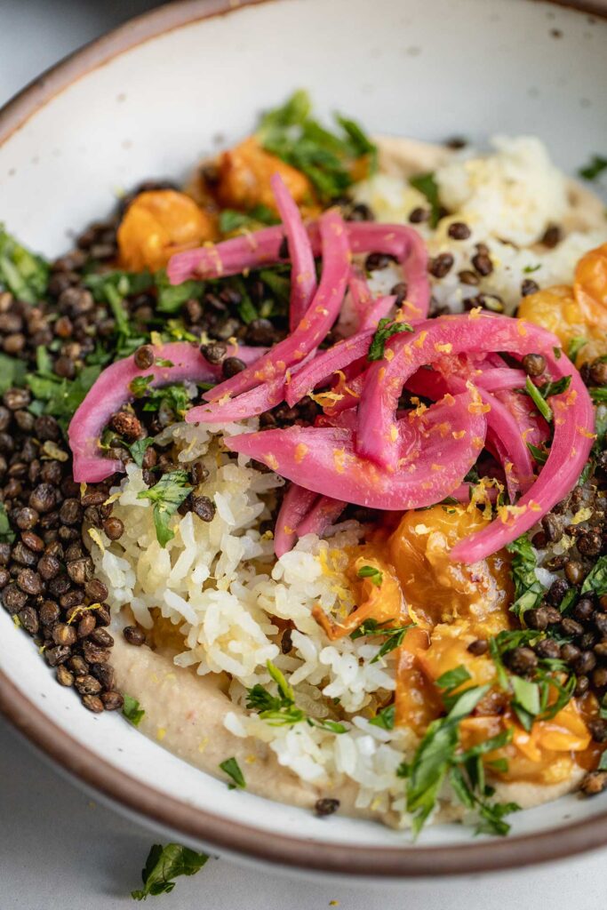 Side view of a bowl of hummus topped with crispy rice, lentils, stewed tomatoes, parsley and pickled onions.