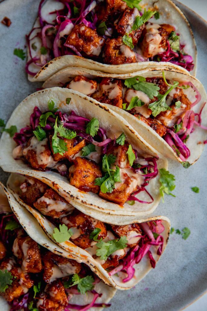 Four tofu tacos stuffed with red cabbage slaw and topped with cilantro lined up on a plate.