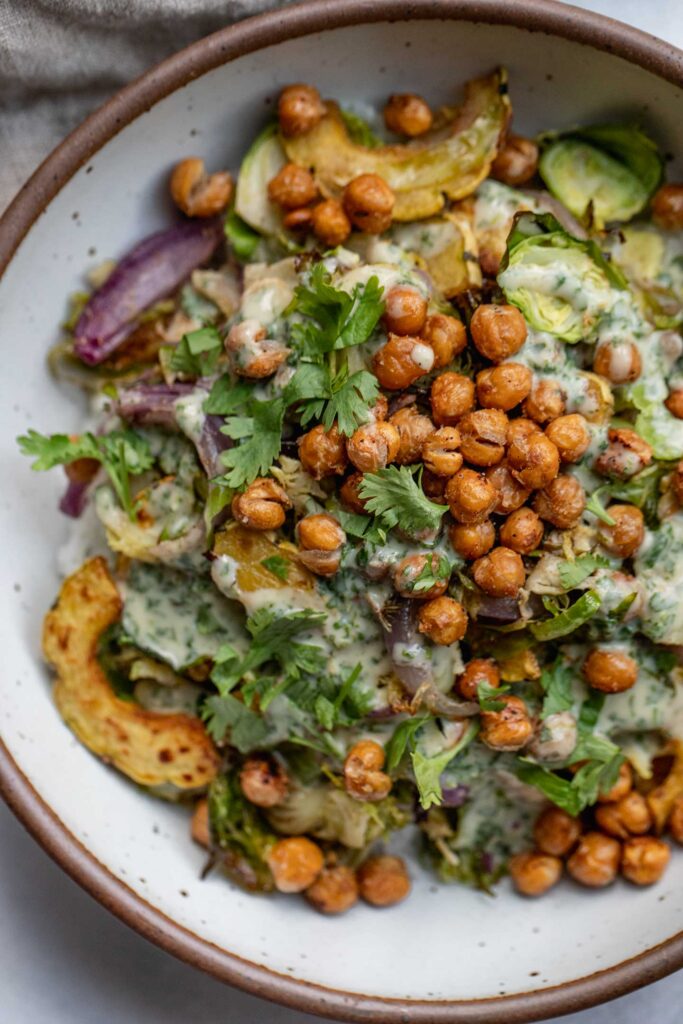 Bowl of roasted squash and brussels sprouts topped with chickpeas and a creamy dressing.