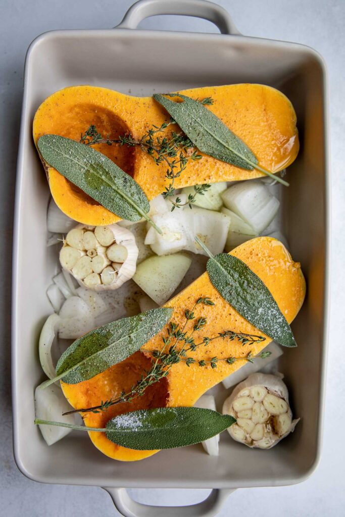 Halved butternut squash, exposed garlic cloves and onion topped with oil, sage and thyme in a baking dish.