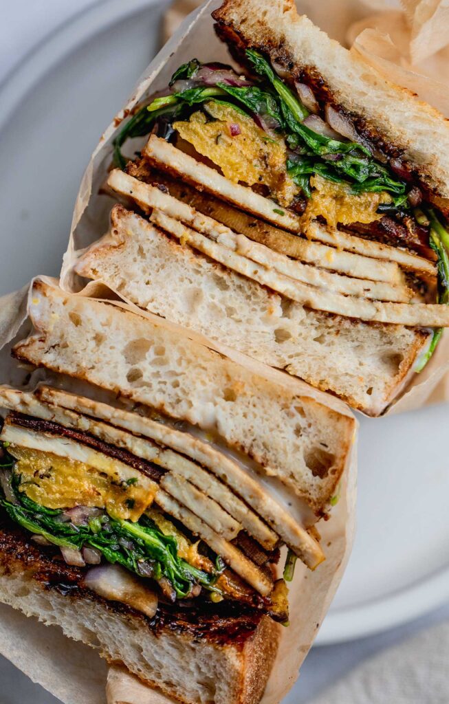 Assembled sandwich, tucked in parchment paper and cut in half to show layers of tofu slices, yogurt sauce, and acorn squash between two slices of bread.