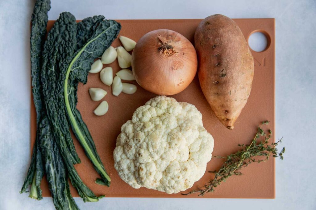 Sweet potato, onion, cauliflower, garlic cloves, kale and thyme on a cutting board.