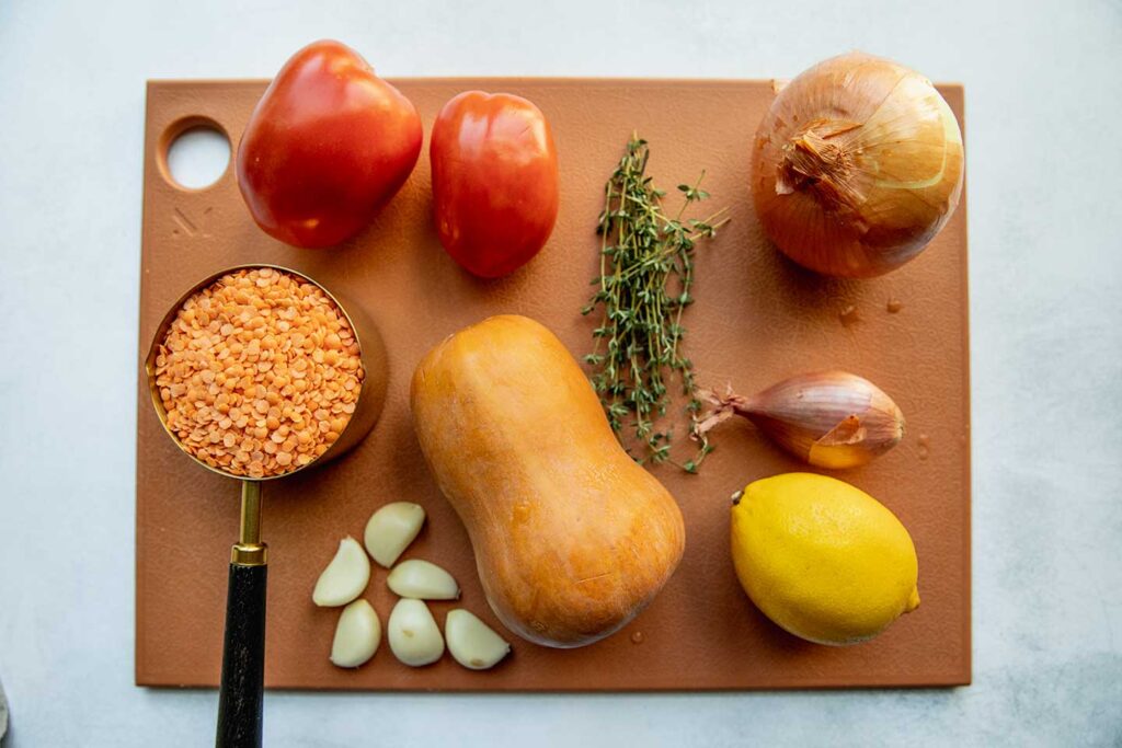 Cutting board topped with plum tomatoes, red lentils, onion, thyme, honeynut squash, shallot, lemon and garlic cloves.