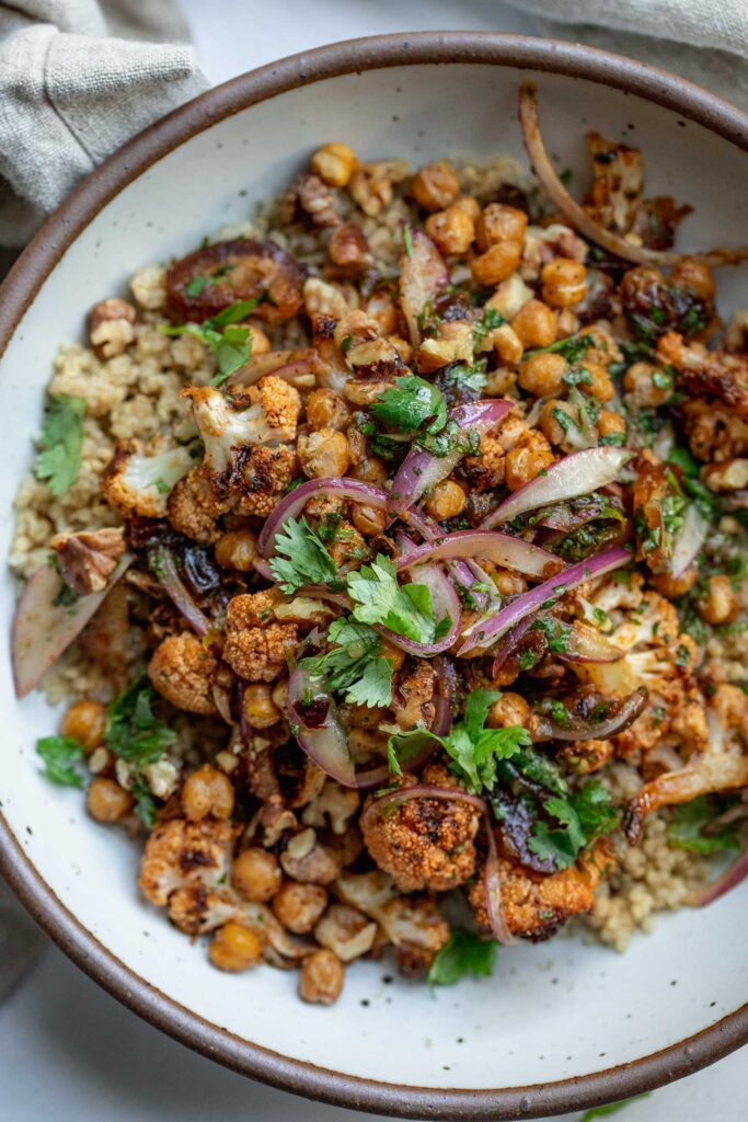 Bowl of quinoa topped with roasted cauliflower tossed with maple lime marinated onions and topped with crispy chickpeas and walnuts.