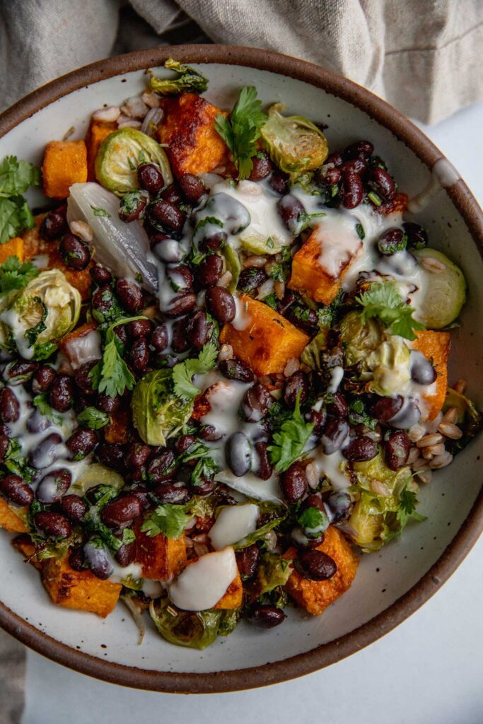 Close up view of a bowl of farro mixed with butternut squash and brussels sprouts topped with marinated black beans and a yogurt sauce.