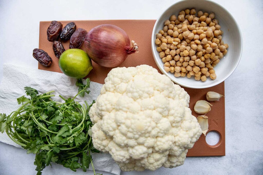 Cutting board topped with dates, lime, onion, cilantro, cauliflower, garlic and chickpeas.