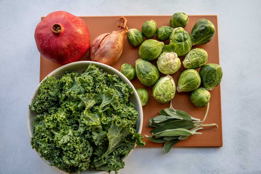 Cutting board topped with brussels sprouts, shallots, kale, sage and pomegranate seeds.