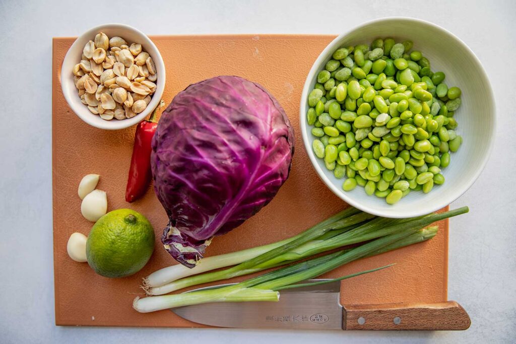 Cutting board with a bowl of peanuts, edamame, cabbage, scallions, lime, garlic and fresno pepper on top.