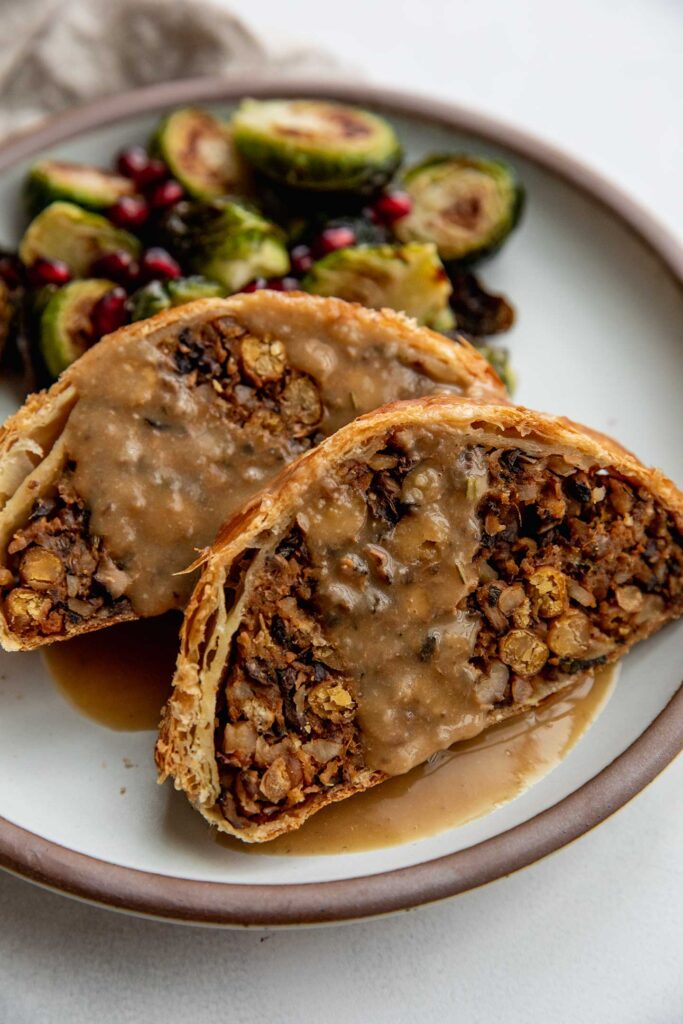 Serving two slices of the vegetable wellington with roasted brussels sprouts on a plate topped with gravy.