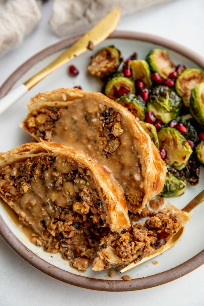 Two slices of the wellington on a plate served with roasted brussels sprouts and pomegrante seeds.