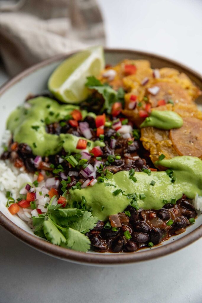 Side view of a bowl of rice topped with chipotle black beans, avocado cilantro sauce and plantains.