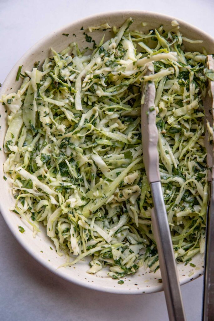 Cabbage slaw and dressing being mixed with tongs in a bowl.