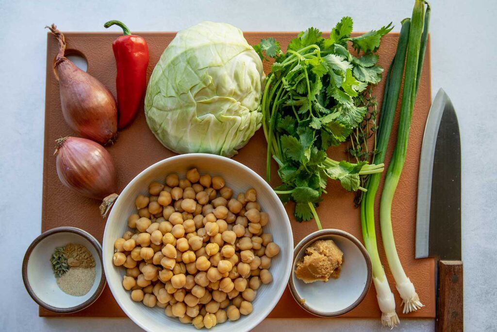 Cutting board topped with cabbage, cilantro, scallions, miso, chickpeas, shallots, pepper and spices.