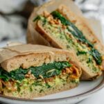 Breakfast sandwich cut in half on a plate showing the layers of avocado, tofu and spinach.