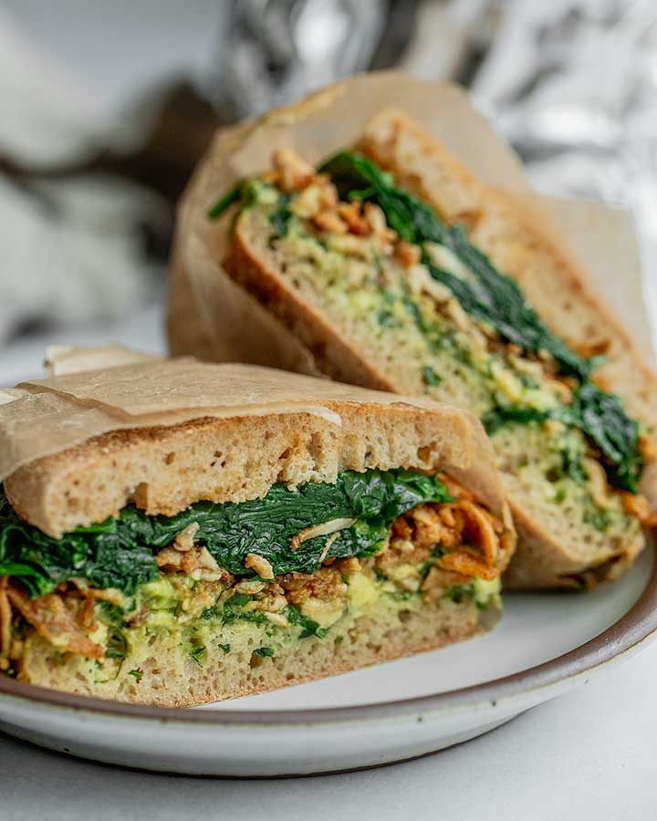 Breakfast sandwich cut in half on a plate showing the layers of avocado, tofu and spinach.