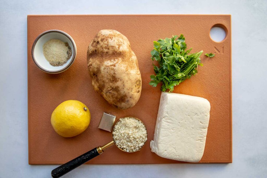 Cutting board topped with tofu, breadcrumbs, lemon, seasonings, potato, bouillon cube and herbs.