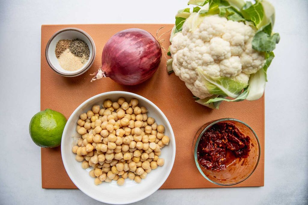Cutting board topped with cauliflower, chipotle peppers, onion, chickpeas, a bowl of seasonings, and limes.