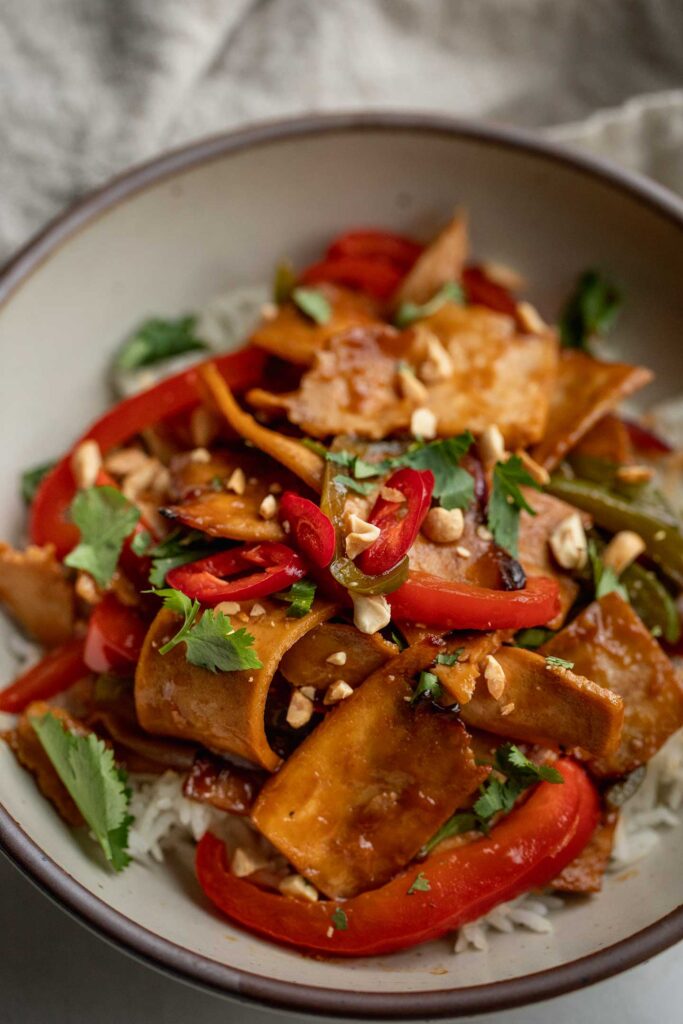 Side view of a bowl of rice topped with pepper and tofu strips, cilantro and peanuts.