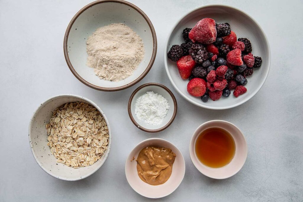 Oats, protein powder, berries, cornstarch, peanut butter, maple syrup on a flat grey surface.