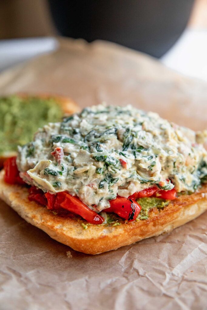 One slice of bread topped with pesto, roasted red bell pepper, and spinach artichoke white bean dip.