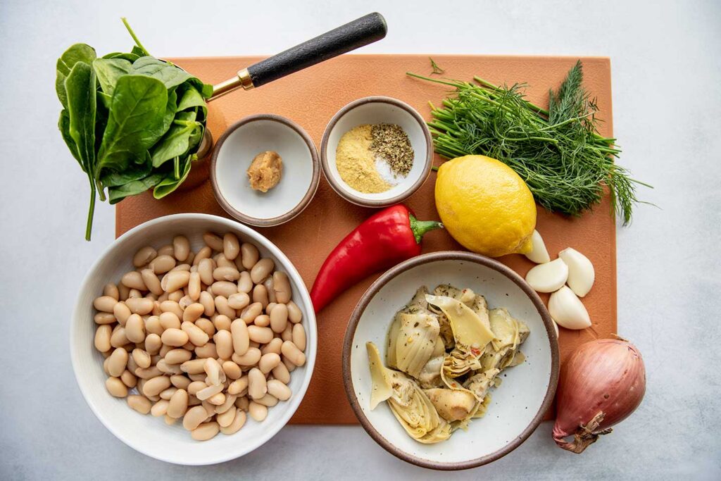 Cutting board topped with spinach, white beans, miso, nutritional yeast, herbs, pepper, lemon, garlic, shallot, and artichoke hearts.