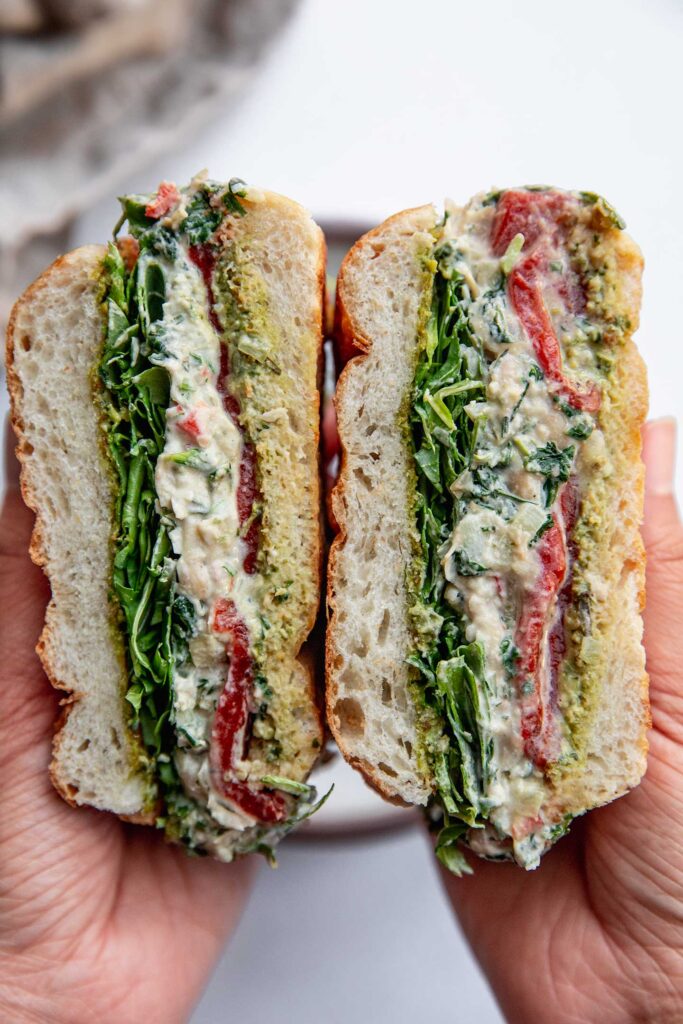 Holding two halves of a cut sandwich stuffed with arugula, spinach artichoke white bean dip, and roasted bell pepper.