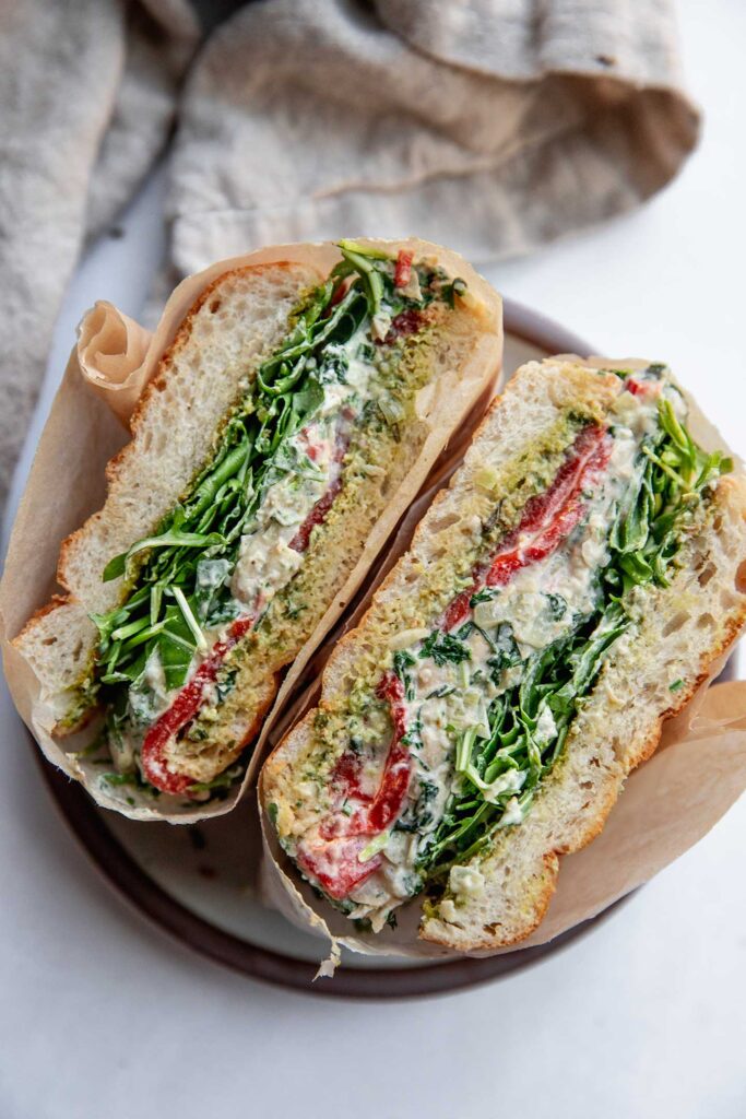 White bean dip sandwich cut in half to show the layered filling of arugula, spinach artichoke white bean dip, and roasted red bell pepper.