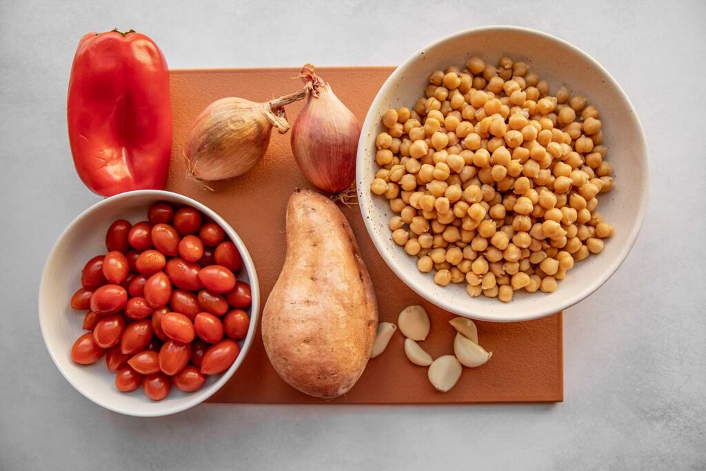 Bell pepper, bowl of chickpeas, shallots, garlic, sweet potato, and cherry tomatoes on a cutting board.