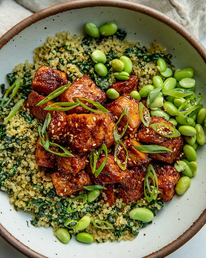 Close up of a bowl of toasted quinoa and kale topped with chili coated tofu, edamame, scallions and sesame seeds.
