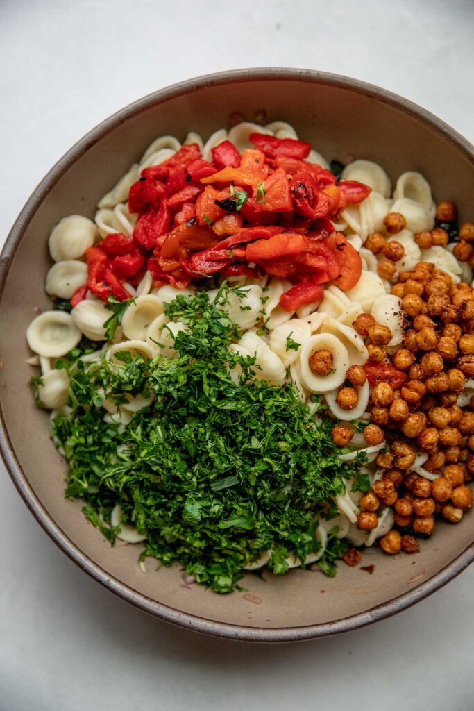 Adding the pasta, sweet peppers, minced herbs and roasted chickpeas in a bowl with kale.