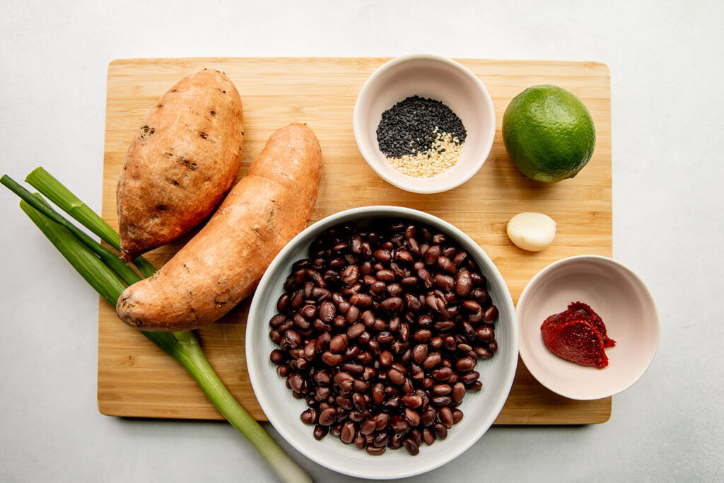 Cutting board with sweet potatoes, black beans, scallions, sesame seeds, lime, garlic and gochujang paste.