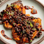 Side view of a plate of baked sweet potato halves loaded with black beans, gochujang sauce and scallions.