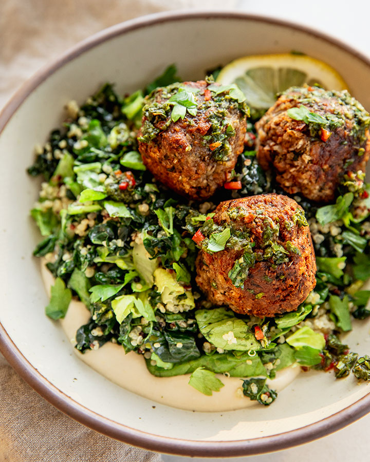 Bowl of white bean spread topped with chimichurri greens and quinoa, chimichurri meatballs and a lemon wedge.