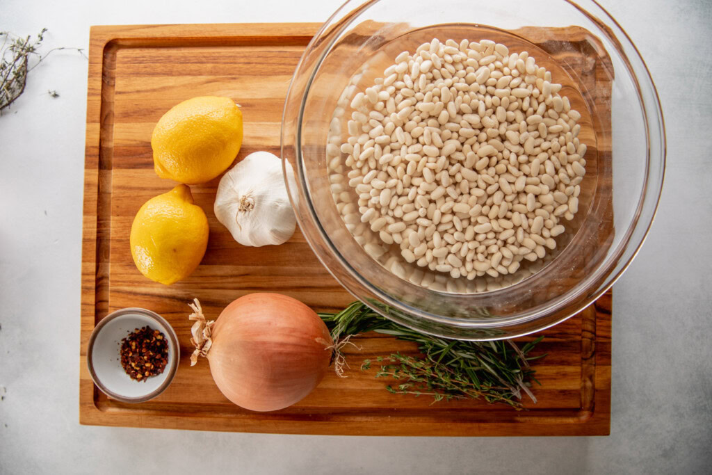 Schneidebrett mit weißen Bohnen, Zwiebeln, Zitrone, Knoblauch, roten Pfefferflocken und Kräutern belegt.