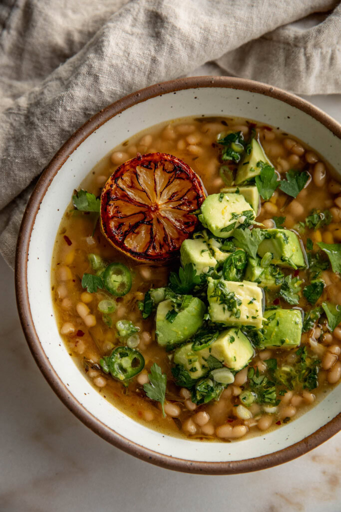 Eine Schüssel mit weißen Bohnen in Brühe, garniert mit Avocado-Salsa und serviert mit einer gerösteten Zitrone.