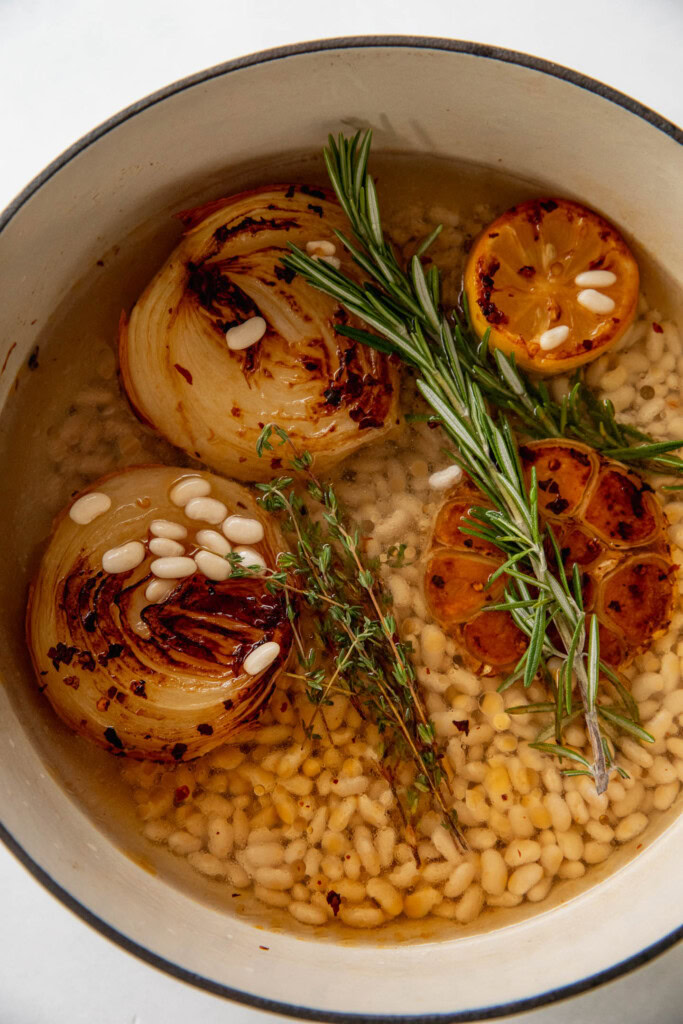 Geben Sie die Bohnen und das Wasser in einen Topf mit gerösteten Zwiebeln, Zitrone, Knoblauch und frischem Rosmarin und Thymian.