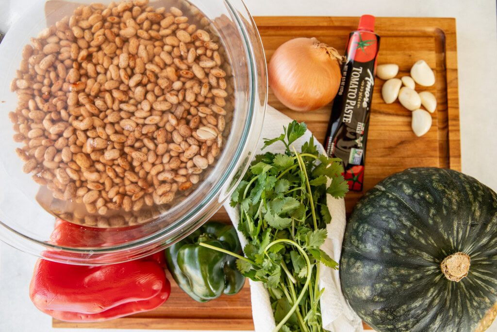 Cutting board with a bowl of soaked pinto beans, bell peppers, kabocha squash, cilantro, onion, tomato paste, and garlic cloves.