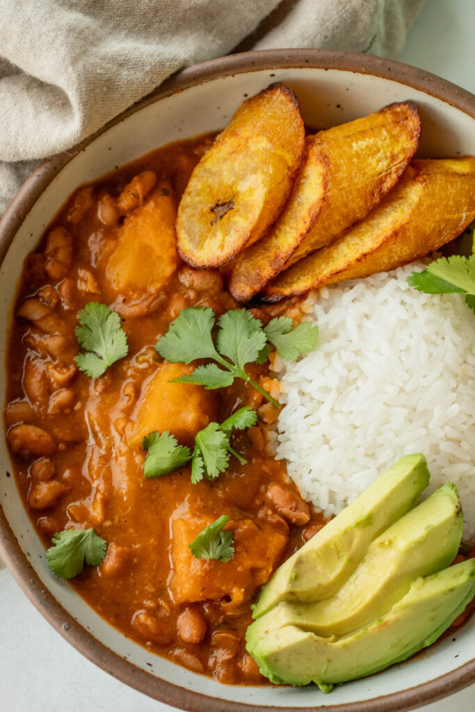 Close up of a bowl of habichuelas guisadas served with a mound of rice, avocado slices, and baked plantains.