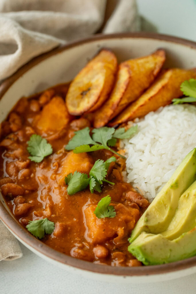 Side view of a bowl of stewed beans served with a mound of rice, avocado slices, and baked plantains.