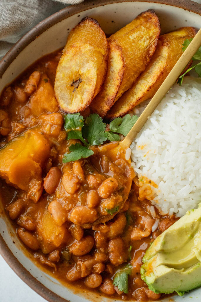 Grabbing a spoonful of stewed beans from a bowl served alongside some rice, plantains and avocado.